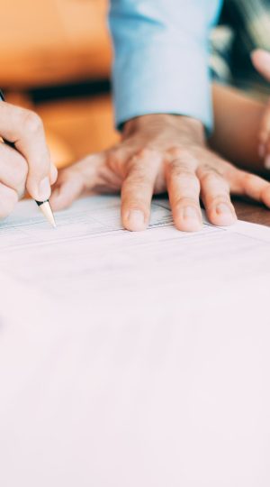 Hand of senior male candidate holding pen and filling in application form together with woman at table. Senior partner signing contract