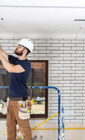 Electrician Builder at work, installation of lamps at height. Professional in overalls with tools on the background of the repair site. Home renovation concept.